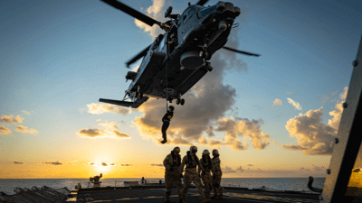 Military personnel on ship with helicopter flying above and soldier dropping being lowered onto deck