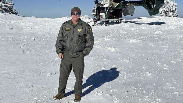 Mike Calhoun posing on snowy mountain with sheriff helicopter behind