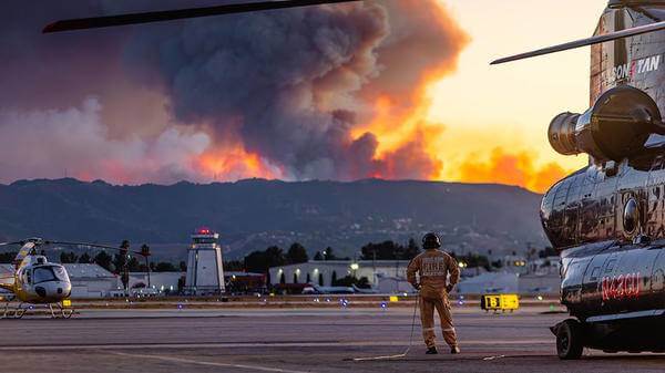 Fire in background of helicopter parked with pilot standing next to it