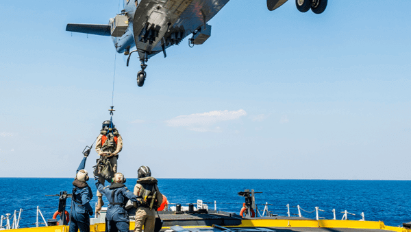Helicopter with rope lowering military personnel onto ship, with military underneath helping unclip